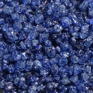 May include: A close-up view of a pile of small, blue, iridescent glass pieces. The glass pieces are irregularly shaped and have a shiny, reflective surface.