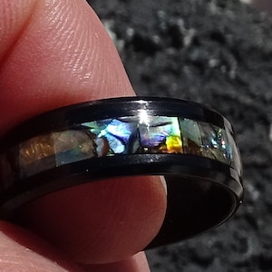 May include: A black ring with a band of iridescent abalone shell inlay. The ring is worn on a finger.