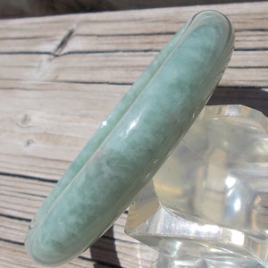 May include: A light green jade bangle bracelet with a smooth, polished finish. The bracelet is displayed on a clear plastic stand.
