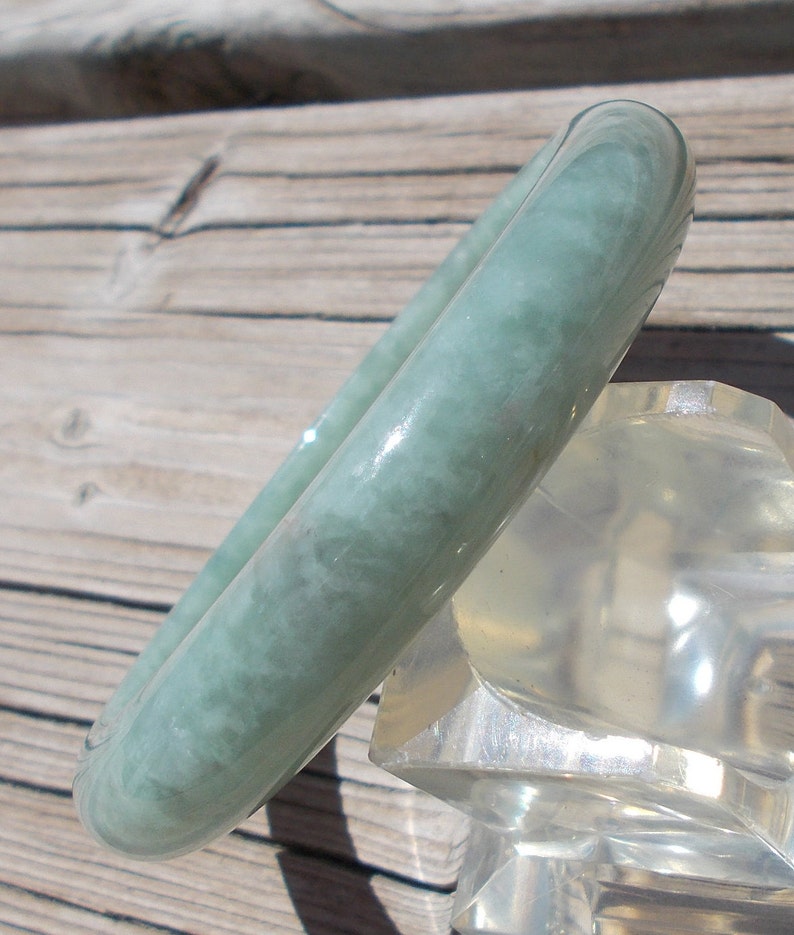 May include: A light green jade bangle bracelet with a smooth, polished finish. The bracelet is displayed on a clear plastic stand.