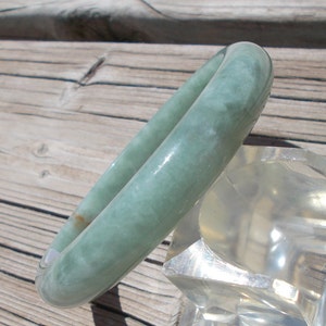 May include: A light green jade bangle bracelet. The bracelet is smooth and polished, with a subtle swirl pattern. It is displayed on a clear stand.