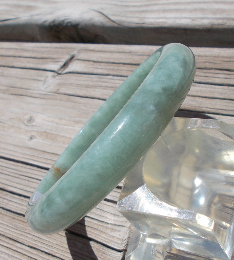 May include: A light green jade bangle bracelet. The bracelet is smooth and polished, with a subtle swirl pattern. It is displayed on a clear stand.