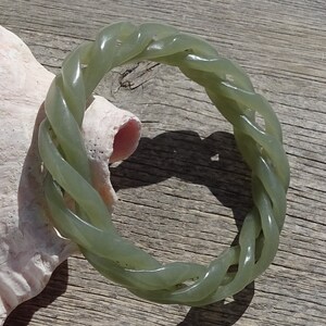 May include: A green jade bangle bracelet with a braided design. The bracelet is made of a smooth, polished jade and has a natural green color.