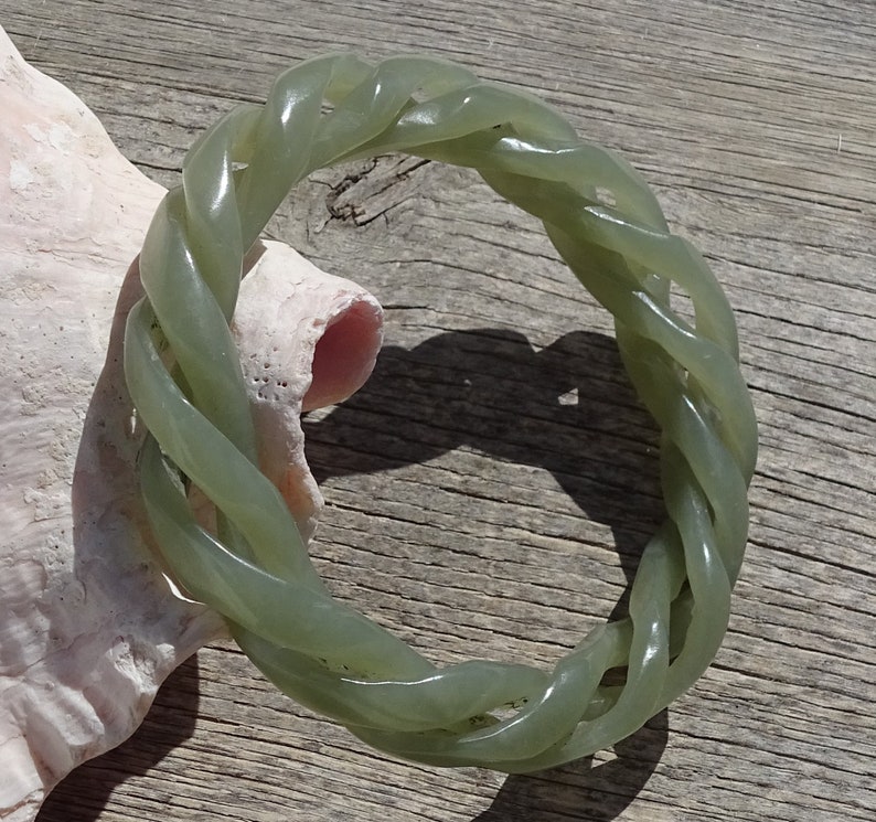 May include: A green jade bangle bracelet with a braided design. The bracelet is made of a smooth, polished jade and has a natural green color.