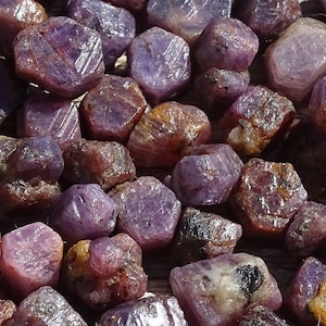 May include: A collection of rough, purple ruby gemstones. The rubies are various sizes and shapes, with some having a more reddish hue.