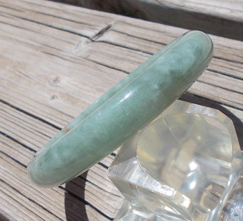 May include: A light green jade bangle bracelet. The bracelet is smooth and polished, with a subtle marbled pattern. It is displayed on a clear plastic stand.