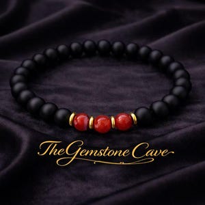 Puede incluir: Una pulsera de cuentas negras con tres cuentas de piedras preciosas rojas y detalles dorados. La pulsera se muestra sobre una superficie oscura y texturizada. El texto "The Gemstone Cave" está en escritura dorada.