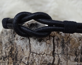 May include: A black leather bracelet with a knotted design. The bracelet is made of two strands of leather that are tied together in a knot.