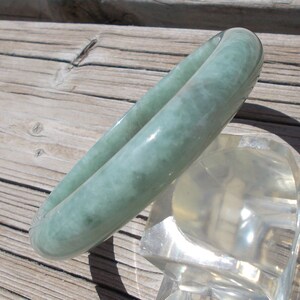 May include: A light green jade bangle bracelet with a cloudy, marbled pattern. The bangle is displayed on a clear plastic stand.
