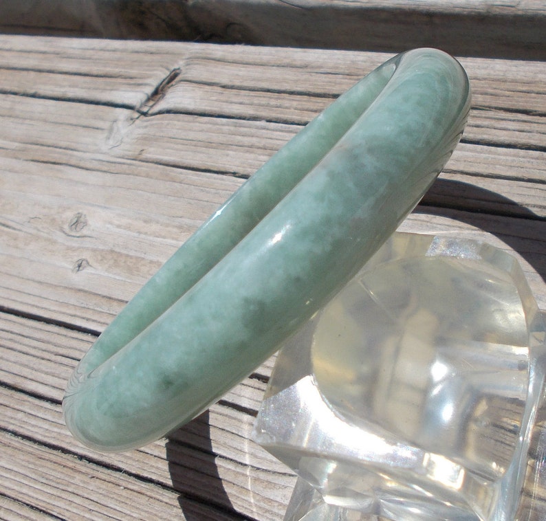 May include: A light green jade bangle bracelet with a cloudy, marbled pattern. The bangle is displayed on a clear plastic stand.