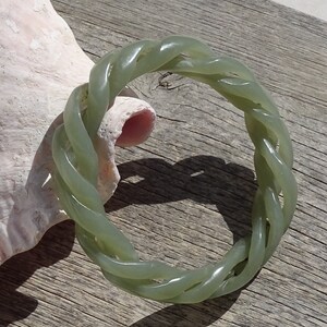 May include: A green jade bangle bracelet with a braided design. The bracelet is made of a smooth, polished jade and has a natural, earthy look.