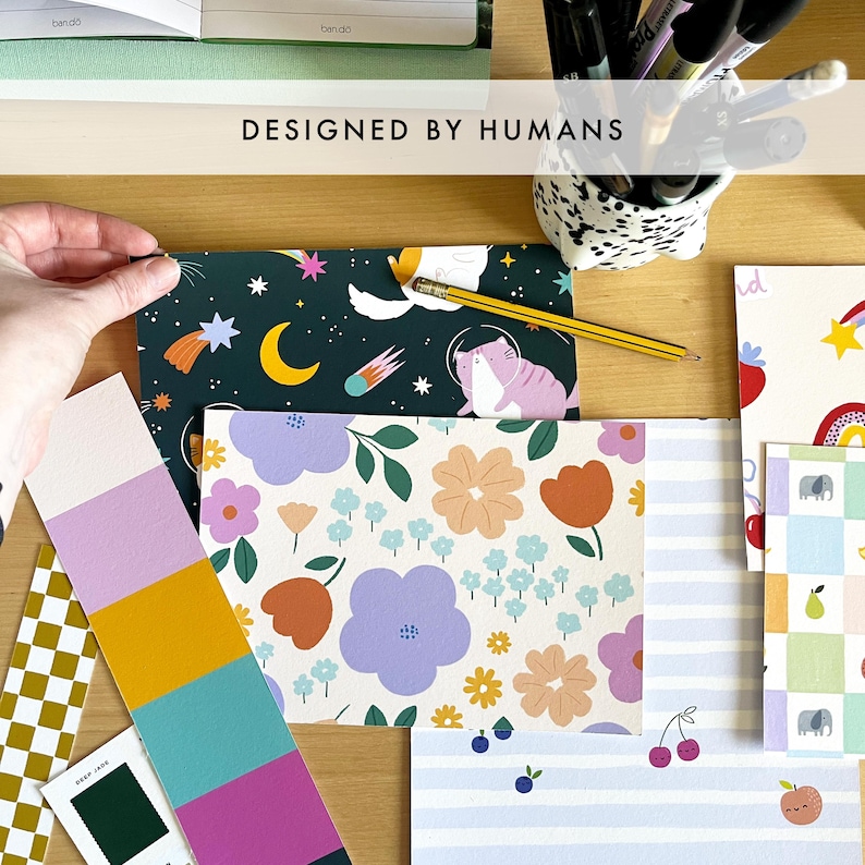 May include: A collection of patterned stationery and color swatches. Designs include floral, celestial, and striped patterns. The text "DESIGNED BY HUMANS" is visible. A hand is holding a piece of stationery.
