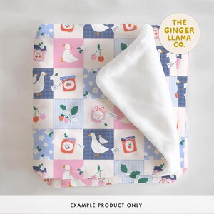 May include: A patchwork baby blanket with a white plush backing. The front features a repeating pattern of geese, jars of jam, cherries, and strawberries in pink, blue, and white squares. The text "THE GINGER LLAMA CO." is in a circle.