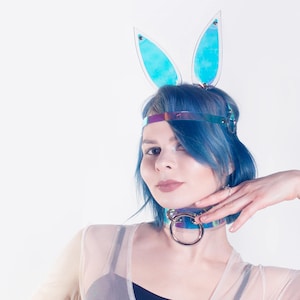 May include: A person with blue hair wearing a holographic headband and a choker with a large ring. The headband has two blue bunny ears attached to it.