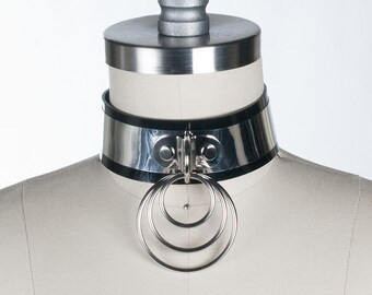 Pode incluir: Um colar choker prateado com uma faixa de couro preta e três anéis entrelaçados. O colar está em um manequim branco.