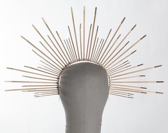 May include: A gold-toned crown with a halo of thin, pointed spikes radiating outward. The crown is displayed on a gray mannequin head.