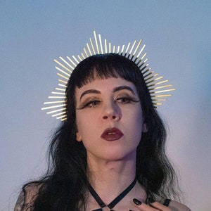 May include: A woman with dark hair and dark makeup wears a gold crown with a pointed design. She is wearing a black lace top with a black harness and has black nail polish.