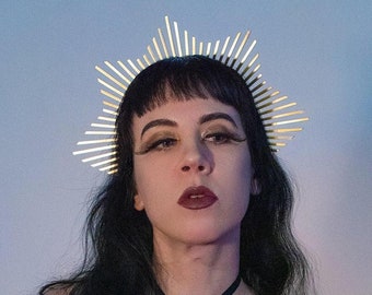 May include: A woman with dark hair and dark makeup wears a gold crown with a pointed design. She is wearing a black lace top with a black harness and has black nail polish.