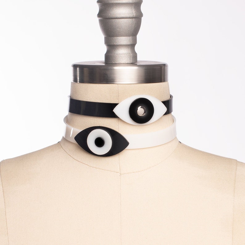 May include: Two black and white eye-shaped chokers on a white mannequin. The top choker is black with a white eye and the bottom choker is white with a black eye.