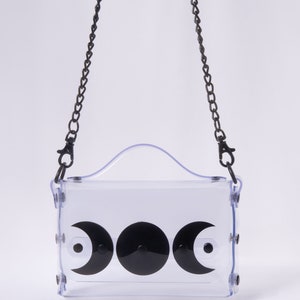 May include: A clear plastic crossbody bag with a black chain strap and three black crescent moon designs. The bag has a top handle.