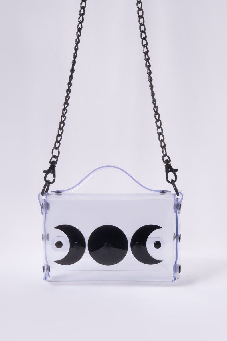 May include: A clear plastic crossbody bag with a black chain strap and three black crescent moon designs. The bag has a top handle.