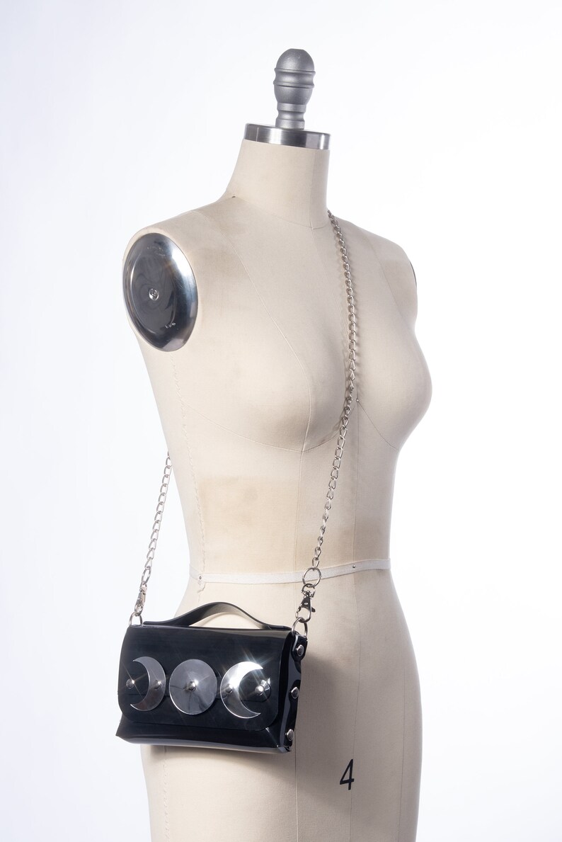 May include: A black patent leather crossbody bag with a silver chain strap and three silver crescent moon embellishments.
