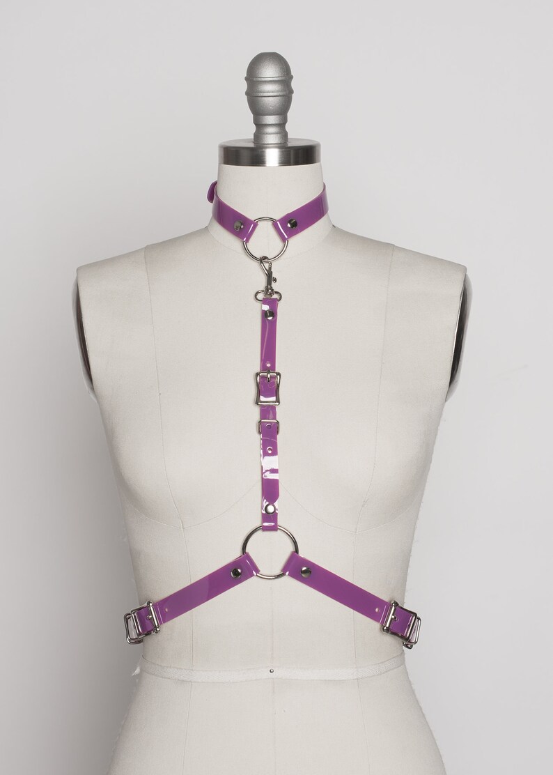 May include: Purple vinyl harness with silver hardware, including a choker, a ring, and a buckle. The harness is on a white mannequin.