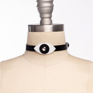 May include: A black choker necklace with a white eye-shaped pendant. The pendant has a black pupil.
