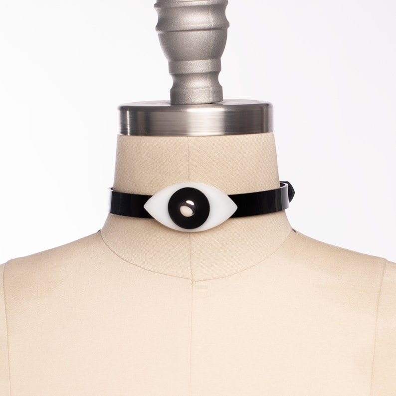 May include: A black choker necklace with a white eye-shaped pendant. The pendant has a black pupil.