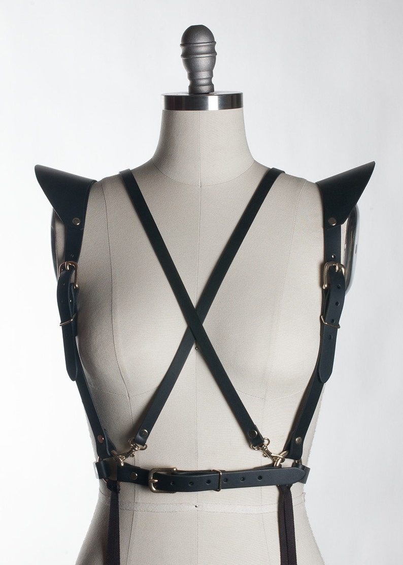 May include: Black leather harness with gold hardware and shoulder straps. The harness has a belt with a buckle and two straps that cross in the front.