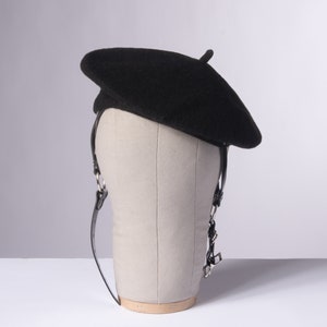 May include: A black beret with a leather strap and silver buckle sits on a gray mannequin head.
