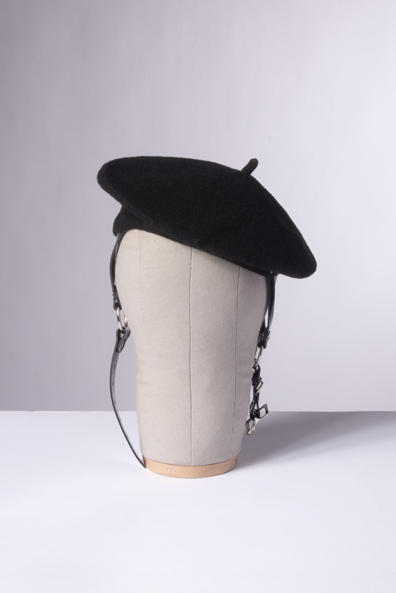 May include: A black beret with a leather strap and silver buckle sits on a gray mannequin head.