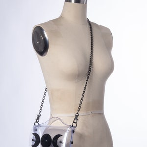 May include: A clear plastic crossbody bag with a black chain strap and three black crescent moon shapes on the front. The bag is on a white mannequin.