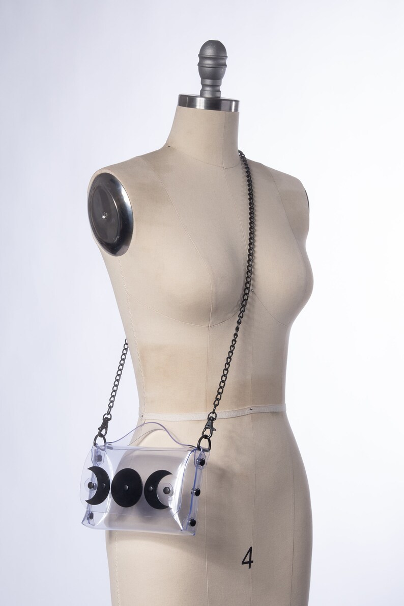 May include: A clear plastic crossbody bag with a black chain strap and three black crescent moon shapes on the front. The bag is on a white mannequin.