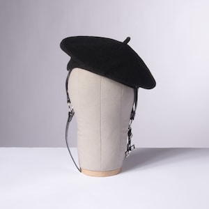 May include: A black beret with a leather strap and silver buckle. The beret is on a mannequin head.