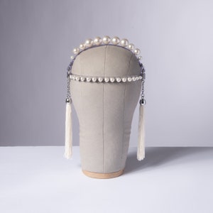 May include: A pearl-adorned headpiece with a clear band and dangling chain tassels. The headpiece features a row of large pearls along the top and a smaller row of pearls at the forehead. The tassels are white and hang down on either side.