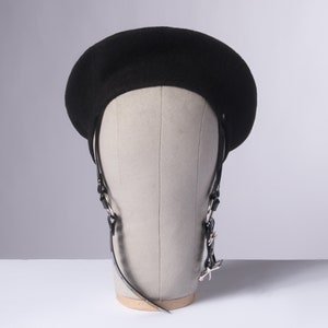 May include: A black beret with a black leather strap and silver hardware. The beret is on a mannequin head.