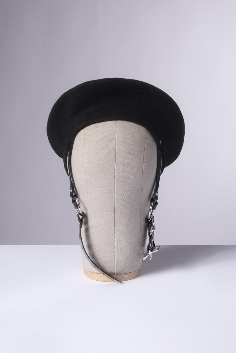 May include: A black beret with a black leather strap and silver hardware. The beret is on a mannequin head.