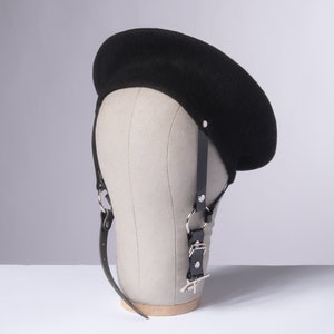 May include: A black beret with a black leather strap and silver hardware. The beret is on a mannequin head.