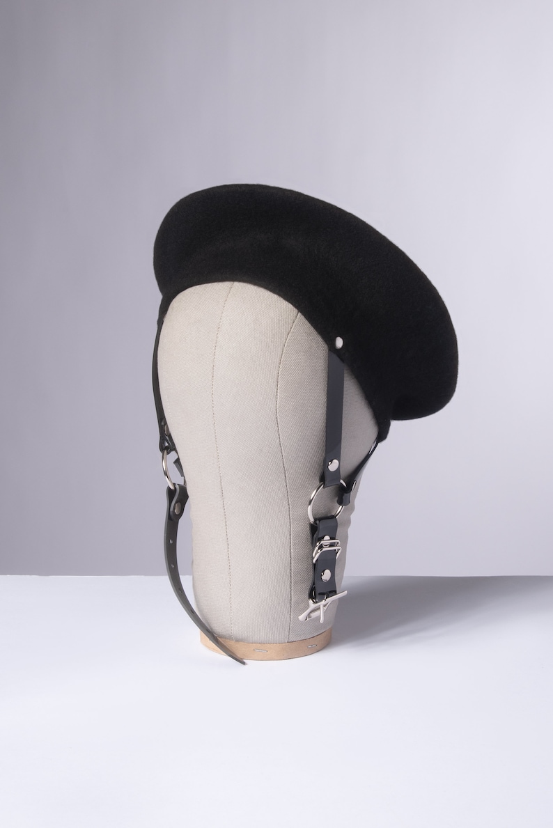 May include: A black beret with a black leather strap and silver hardware. The beret is on a mannequin head.