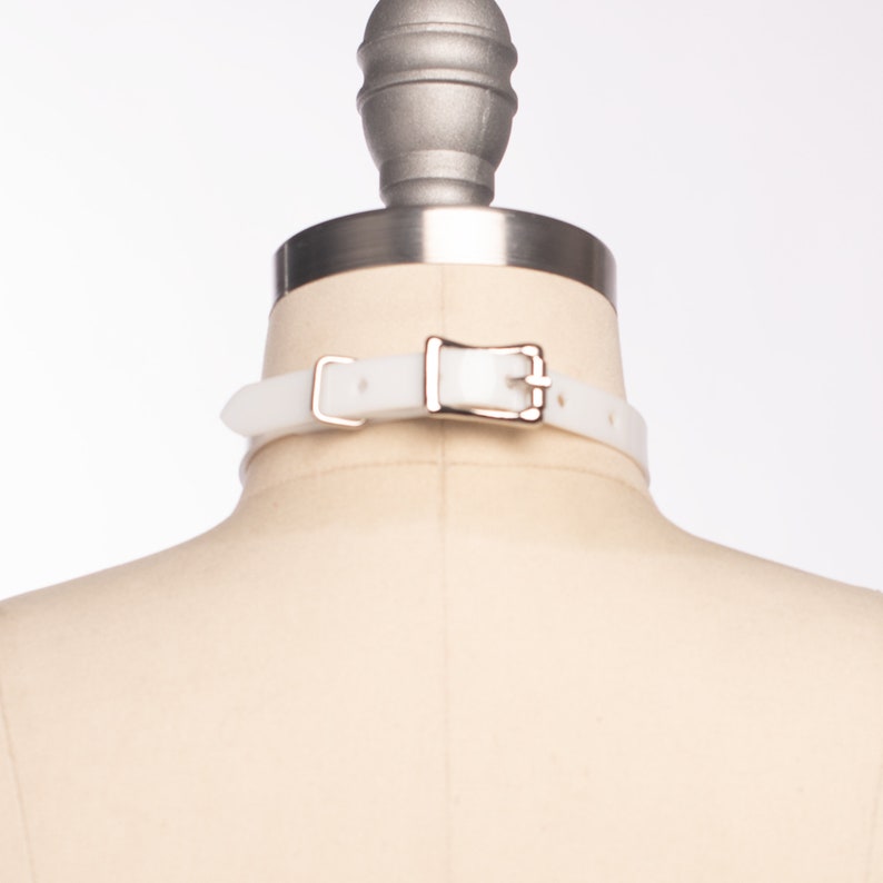 May include: White rubber choker with a silver buckle. The choker is on a white mannequin.
