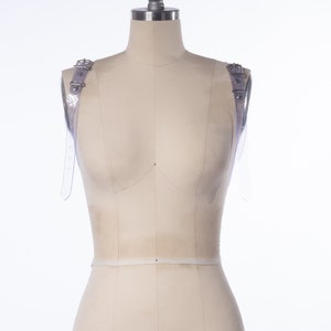 Puede incluir: Un maniqu&iacute; blanco con correas de pl&aacute;stico transparente en cada hombro. Las correas tienen hebillas plateadas. El maniqu&iacute; est&aacute; cubierto con una tela blanca y tiene el n&uacute;mero "4" impreso en la parte delantera.
