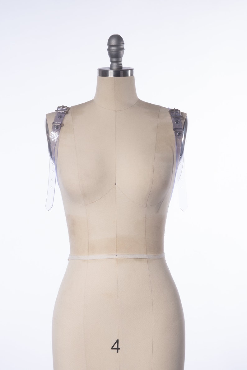 Puede incluir: Un maniqu&iacute; blanco con correas de pl&aacute;stico transparente en cada hombro. Las correas tienen hebillas plateadas. El maniqu&iacute; est&aacute; cubierto con una tela blanca y tiene el n&uacute;mero "4" impreso en la parte delantera.