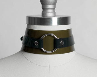 Könnte beinhalten: Ein dunkelgrüner und schwarzer Choker mit einem silbernen Ringakzent. Der Choker ist aus einem glatten, glänzenden Material gefertigt.