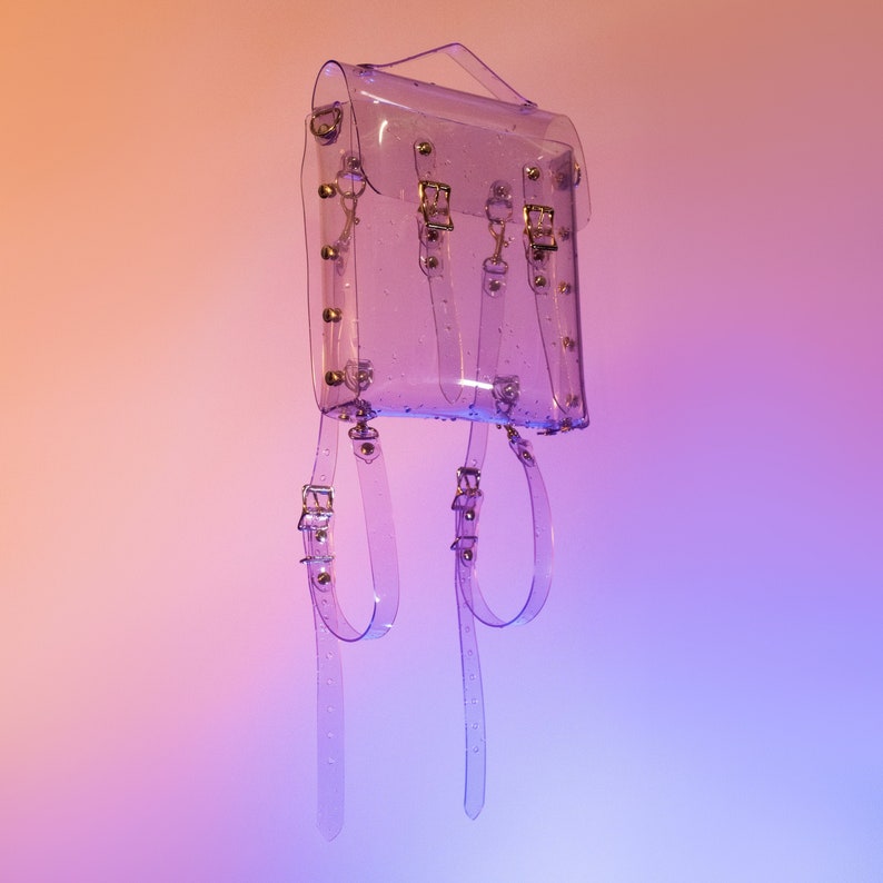 Puede incluir: Una mochila transparente de pl&aacute;stico con detalles dorados y tachuelas. La mochila tiene dos correas ajustables y un asa superior.