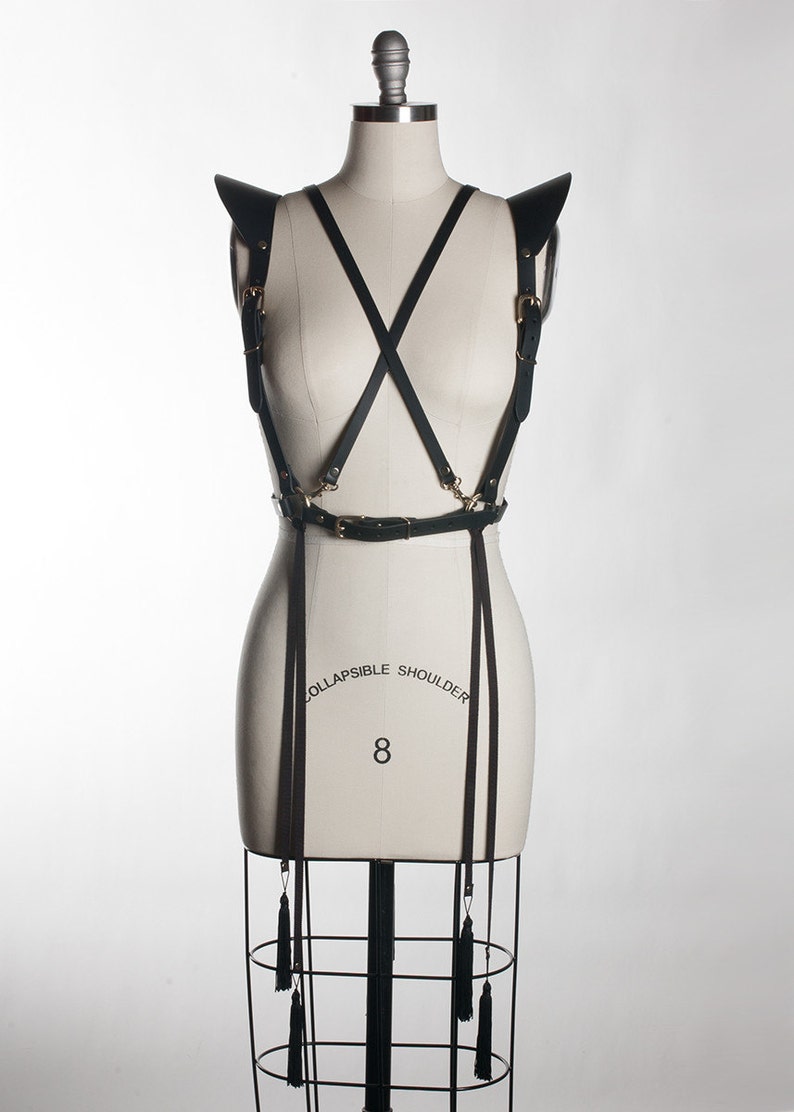 May include: Black leather harness with gold hardware and tassels. The harness has a wide belt and straps that crisscross the chest and back. The straps are adjustable and have buckles. The harness is designed to be worn over the shoulders and chest. The harness is made of high-quality leather and is durable and stylish.