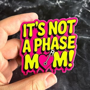 Pode incluir: Um autocolante rosa brilhante com o texto "IT'S NOT A PHASE MOM!" em amarelo, contornado a preto. O design inclui um coração rosa perfurado por um alfinete de segurança prateado. O autocolante tem um efeito de gotejamento.