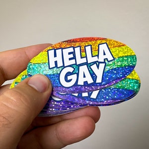 May include: A stack of rainbow-colored, glittery stickers with the text "HELLA GAY" in white letters.