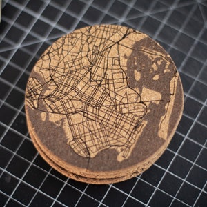 May include: A round cork coaster with a laser-etched map of a city. The coaster is brown and has a natural cork texture.