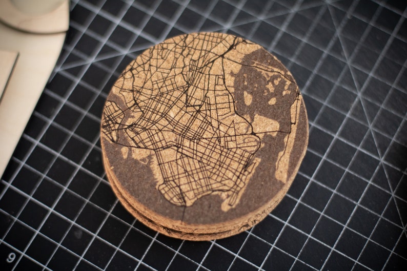 May include: A round cork coaster with a laser-etched map of a city. The coaster is brown and has a natural cork texture.